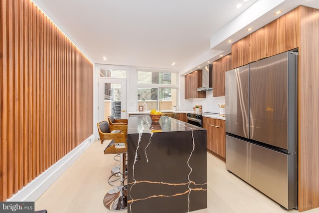 kitchen featuring a center island, brown cabinets, freestanding refrigerator, wall chimney range hood, and modern cabinets