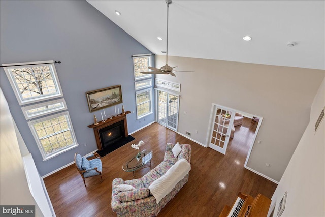 living area featuring high vaulted ceiling, a glass covered fireplace, wood finished floors, baseboards, and ceiling fan