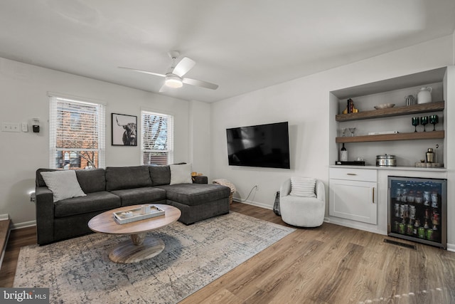 living area with light wood-style flooring, a ceiling fan, a bar, beverage cooler, and baseboards