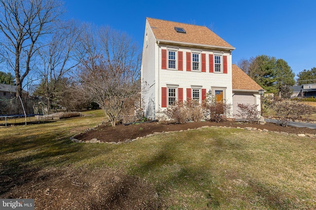 colonial-style house with an attached garage, a front yard, a trampoline, and a shingled roof