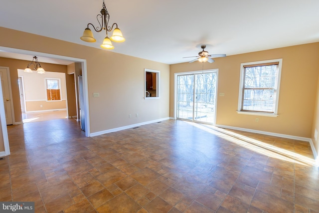 empty room featuring stone finish floor, a healthy amount of sunlight, ceiling fan with notable chandelier, and baseboards