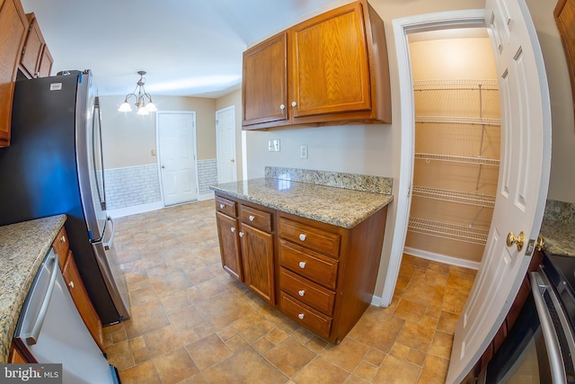 kitchen featuring brown cabinets, stone finish flooring, freestanding refrigerator, wainscoting, and a chandelier