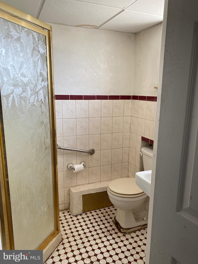 bathroom with a drop ceiling, tile walls, toilet, and a shower with door