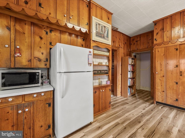 kitchen with stainless steel microwave, freestanding refrigerator, light countertops, wood walls, and light wood-style floors