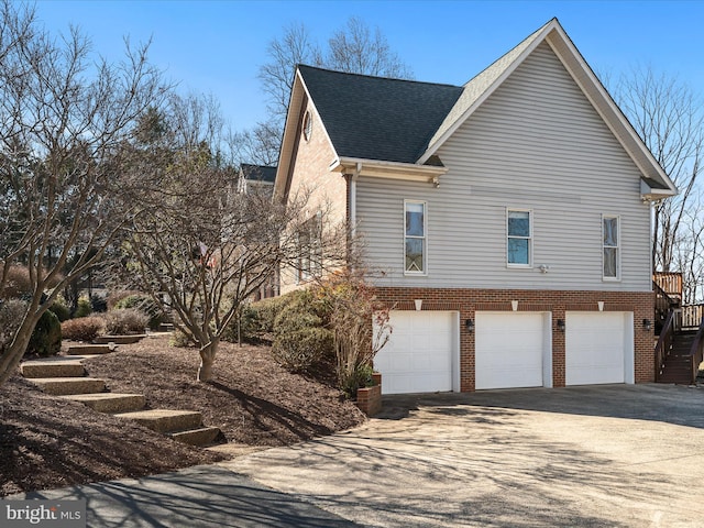 view of side of home with an attached garage, brick siding, stairs, driveway, and roof with shingles