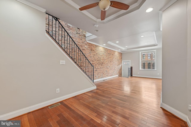 unfurnished living room with a tray ceiling, wood-type flooring, visible vents, and brick wall