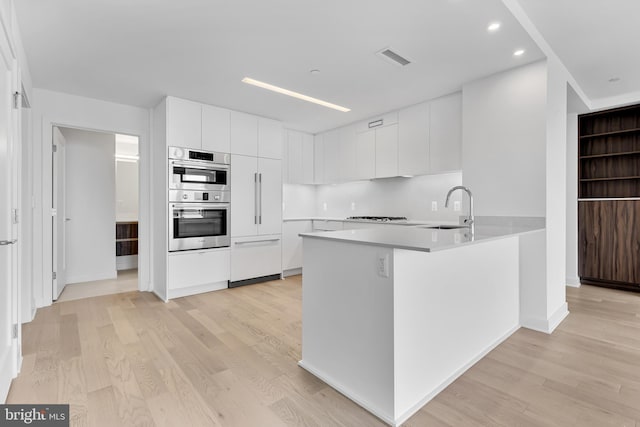 kitchen with white cabinetry, kitchen peninsula, stainless steel double oven, light hardwood / wood-style floors, and sink