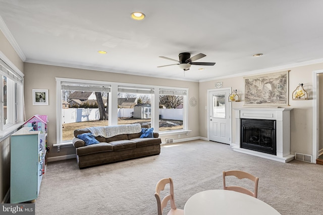 carpeted living room featuring a ceiling fan, visible vents, baseboards, ornamental molding, and a glass covered fireplace