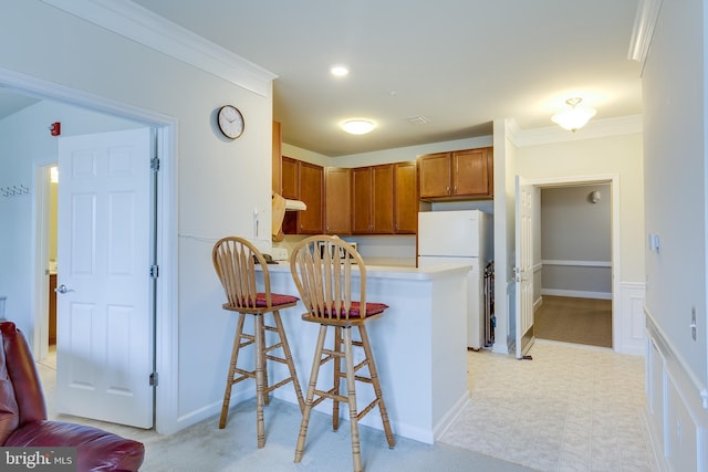 kitchen featuring brown cabinetry, a kitchen breakfast bar, freestanding refrigerator, a peninsula, and crown molding