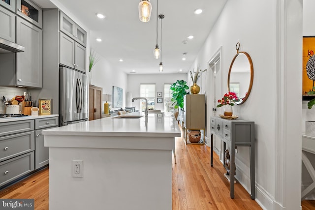 kitchen featuring glass insert cabinets, stainless steel refrigerator, hanging light fixtures, a kitchen island with sink, and light countertops