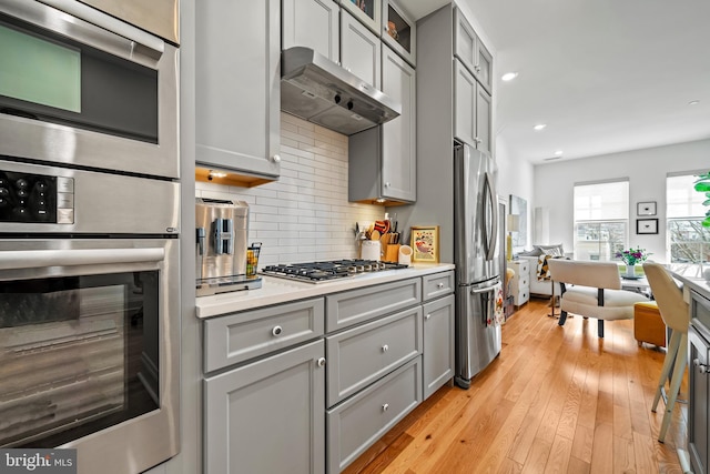 kitchen featuring under cabinet range hood, light countertops, appliances with stainless steel finishes, decorative backsplash, and glass insert cabinets