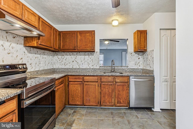 kitchen featuring stone counters, under cabinet range hood, stainless steel appliances, a sink, and brown cabinetry