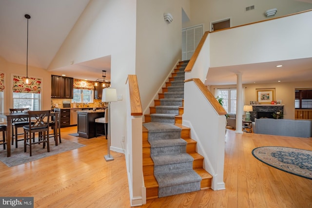 staircase with visible vents, high vaulted ceiling, wood finished floors, a stone fireplace, and decorative columns