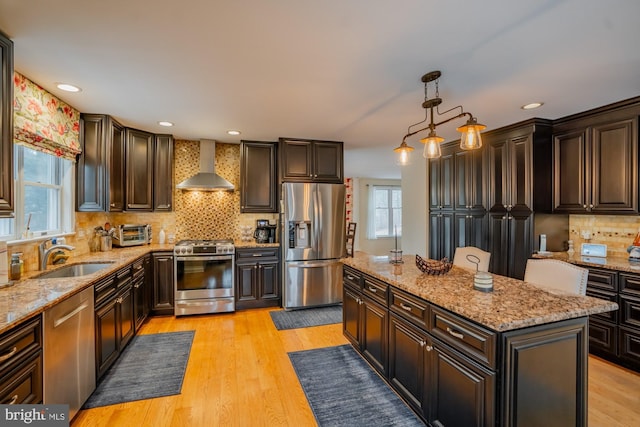 kitchen featuring light wood finished floors, a kitchen island, a sink, stainless steel appliances, and wall chimney exhaust hood