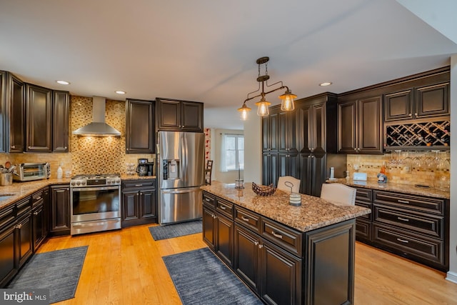 kitchen with a center island, stainless steel appliances, light wood-style floors, wall chimney range hood, and hanging light fixtures