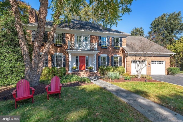 colonial home with aphalt driveway, an attached garage, a balcony, brick siding, and a front yard