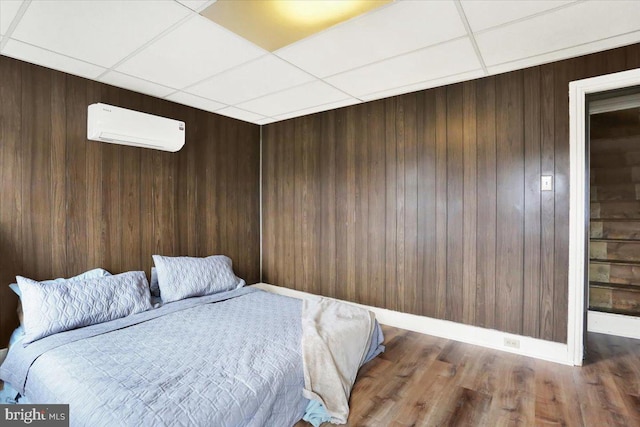 unfurnished bedroom with a paneled ceiling, wood walls, wood finished floors, baseboards, and an AC wall unit