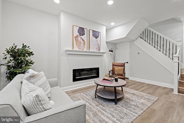 living room featuring a glass covered fireplace, wood finished floors, recessed lighting, baseboards, and stairs