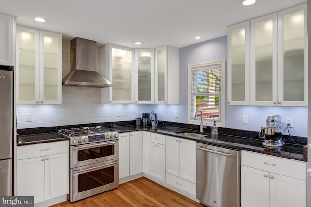 kitchen with wall chimney exhaust hood, stainless steel appliances, sink, white cabinetry, and dark stone countertops