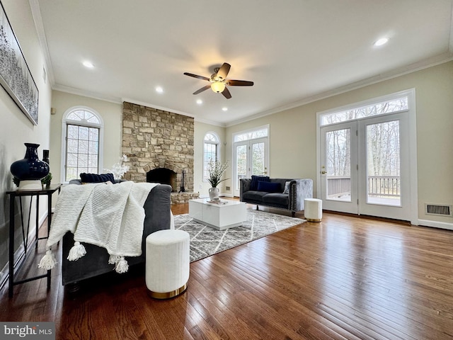 living area featuring ceiling fan, a stone fireplace, wood finished floors, visible vents, and crown molding