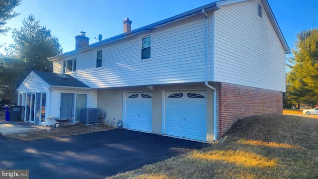 rear view of house featuring brick siding, central AC unit, a chimney, driveway, and an attached garage