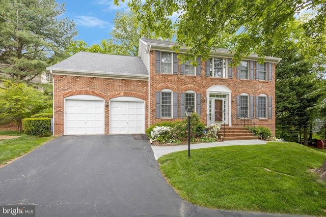 colonial inspired home featuring aphalt driveway, brick siding, roof with shingles, an attached garage, and a front yard