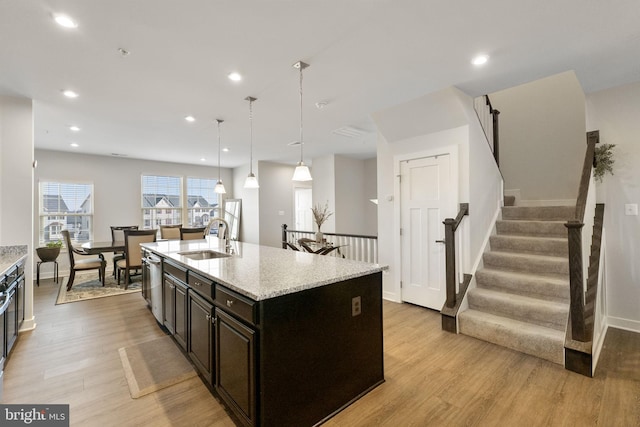 kitchen with light stone counters, light wood finished floors, a kitchen island with sink, a sink, and dishwasher