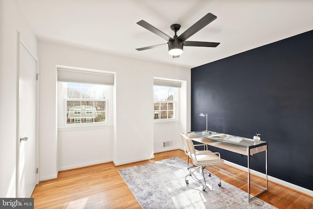 office with an accent wall, wood finished floors, a ceiling fan, visible vents, and baseboards