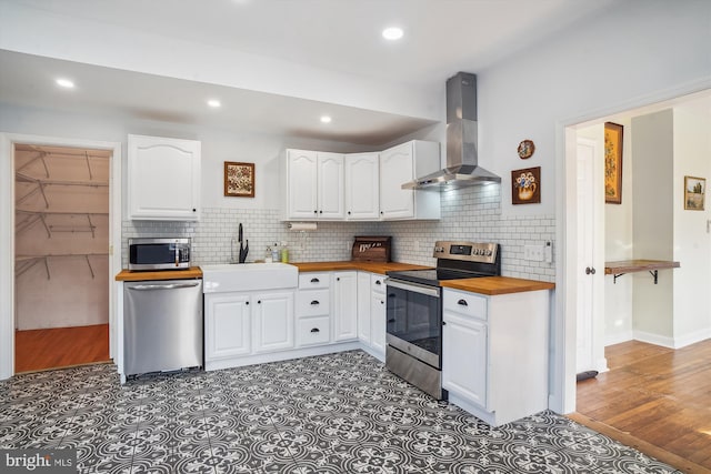 kitchen with butcher block counters, wall chimney exhaust hood, stainless steel appliances, sink, and white cabinetry