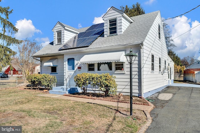 cape cod house with solar panels, a shingled roof, a front yard, and fence