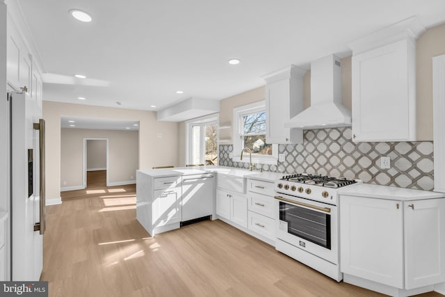 kitchen with a peninsula, white appliances, a sink, white cabinets, and wall chimney exhaust hood