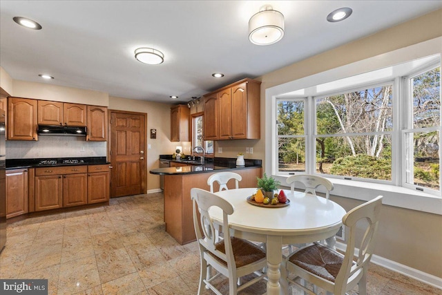 kitchen with under cabinet range hood, black cooktop, a sink, baseboards, and brown cabinets