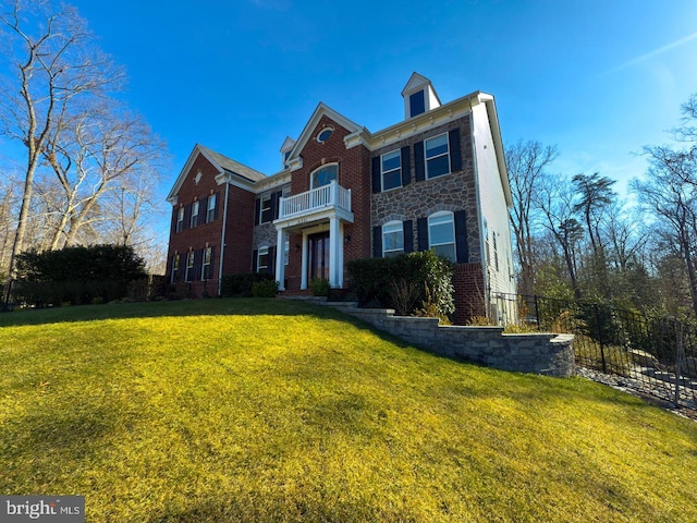 georgian-style home with brick siding, a front lawn, a balcony, and fence