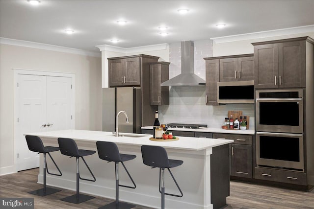 kitchen featuring a kitchen island with sink, ornamental molding, a sink, stainless steel appliances, and wall chimney exhaust hood