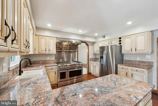 kitchen featuring wall chimney exhaust hood, cream cabinets, appliances with stainless steel finishes, and a sink