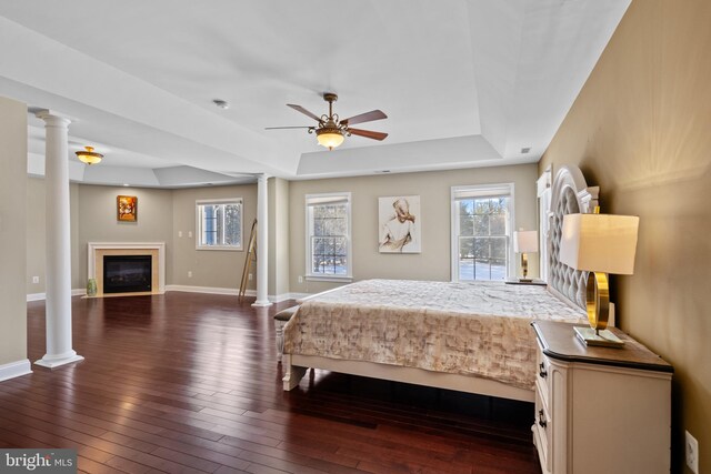 bedroom featuring dark wood-style floors, a tray ceiling, a glass covered fireplace, and ornate columns