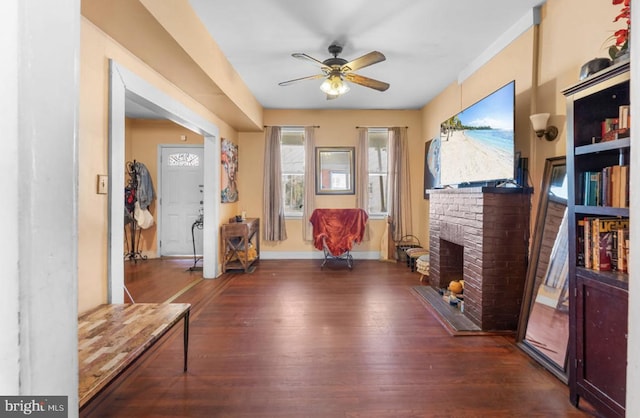 sitting room featuring dark wood-type flooring, a brick fireplace, ceiling fan, and baseboards