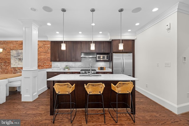 kitchen featuring appliances with stainless steel finishes, hanging light fixtures, dark brown cabinets, an island with sink, and decorative columns