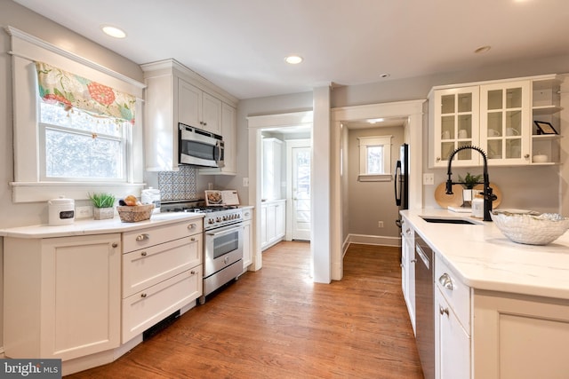 kitchen with stainless steel appliances, a sink, light wood finished floors, glass insert cabinets, and tasteful backsplash