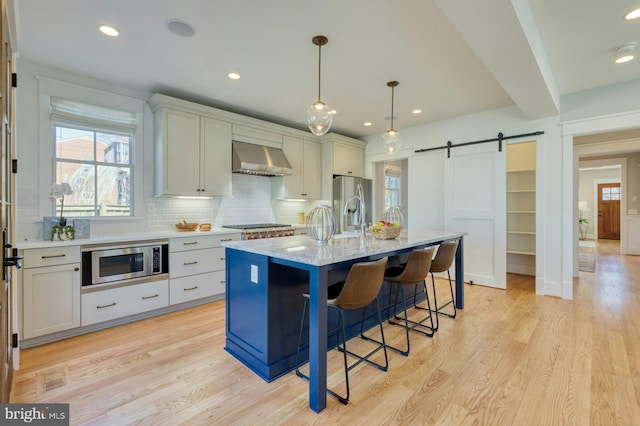 kitchen featuring wall chimney range hood, a barn door, appliances with stainless steel finishes, and light wood finished floors