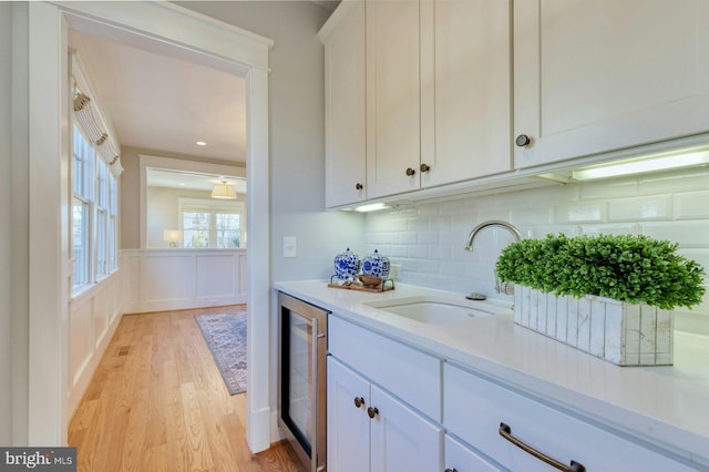 bar featuring beverage cooler, a wainscoted wall, a sink, light wood-type flooring, and tasteful backsplash