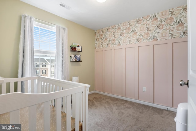 bedroom featuring visible vents, a wainscoted wall, a nursery area, wallpapered walls, and light colored carpet