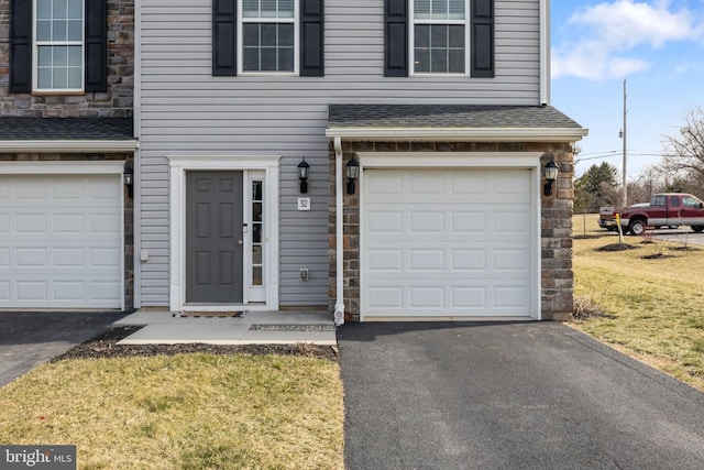 view of exterior entry with a yard, a shingled roof, a garage, stone siding, and aphalt driveway