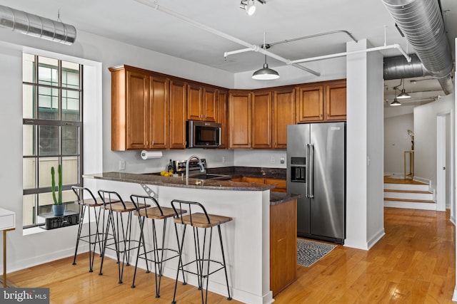 kitchen featuring appliances with stainless steel finishes, brown cabinetry, dark stone countertops, light wood-type flooring, and a peninsula