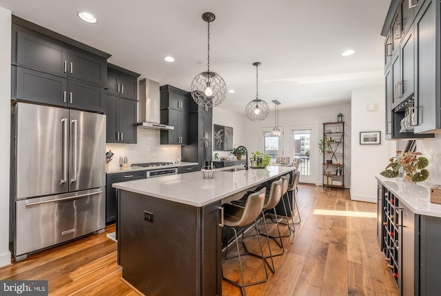 kitchen featuring decorative light fixtures, wall chimney exhaust hood, stainless steel appliances, an island with sink, and a kitchen breakfast bar