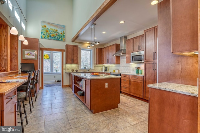 kitchen with tasteful backsplash, stainless steel microwave, a kitchen island, wall chimney range hood, and brown cabinets