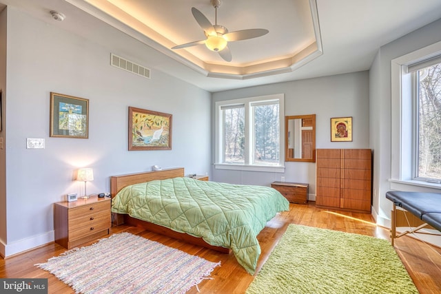 bedroom with light wood-type flooring, visible vents, a ceiling fan, baseboards, and a raised ceiling