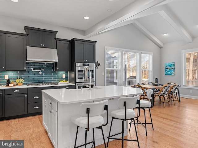 kitchen featuring vaulted ceiling with beams, a breakfast bar area, stainless steel appliances, light countertops, and an island with sink