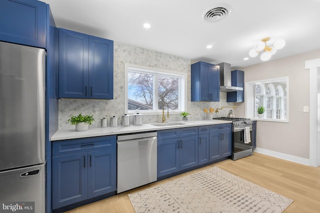 kitchen with appliances with stainless steel finishes, wall chimney exhaust hood, blue cabinetry, light wood-type flooring, and sink