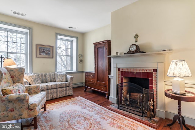 living area featuring a tiled fireplace, wood finished floors, visible vents, and baseboard heating